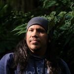 A contemplative man with long hair stands in a forest, bathed in filtered sunlight.